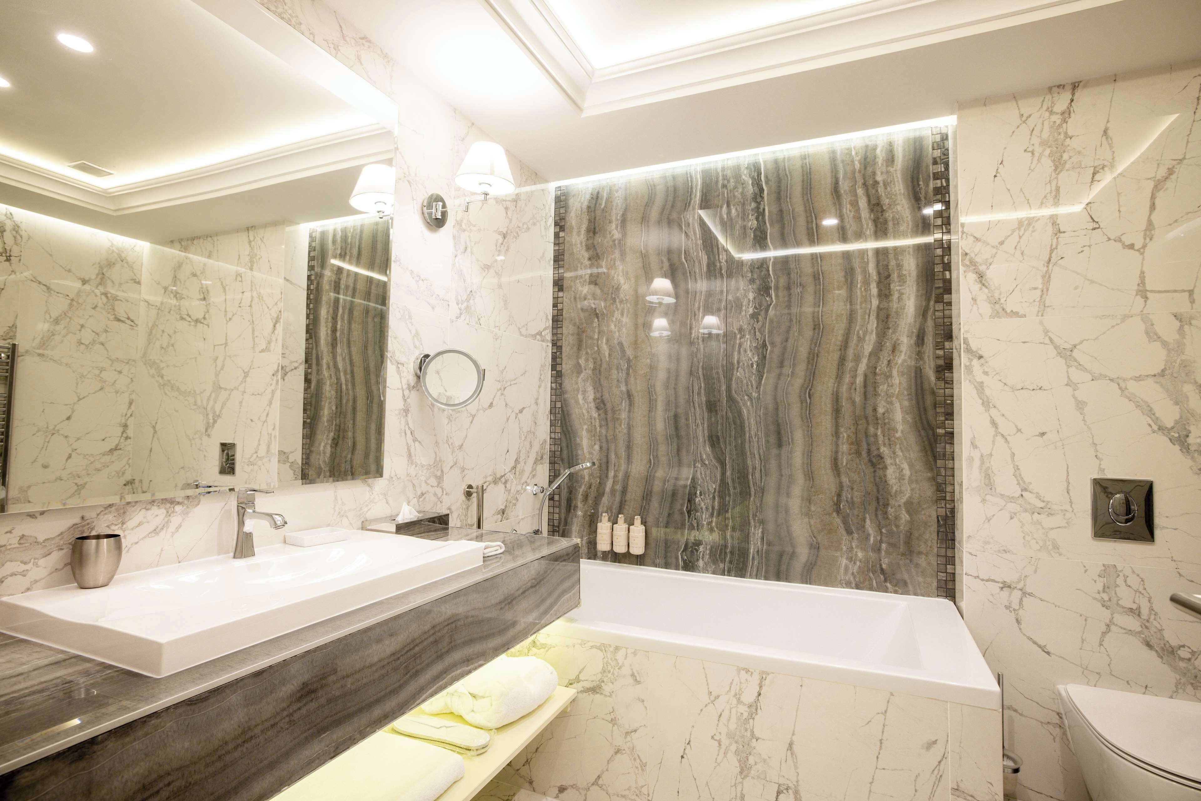 Un baño moderno con un espejo grande, dos lavamanos, una bañera y paredes de mármol.
