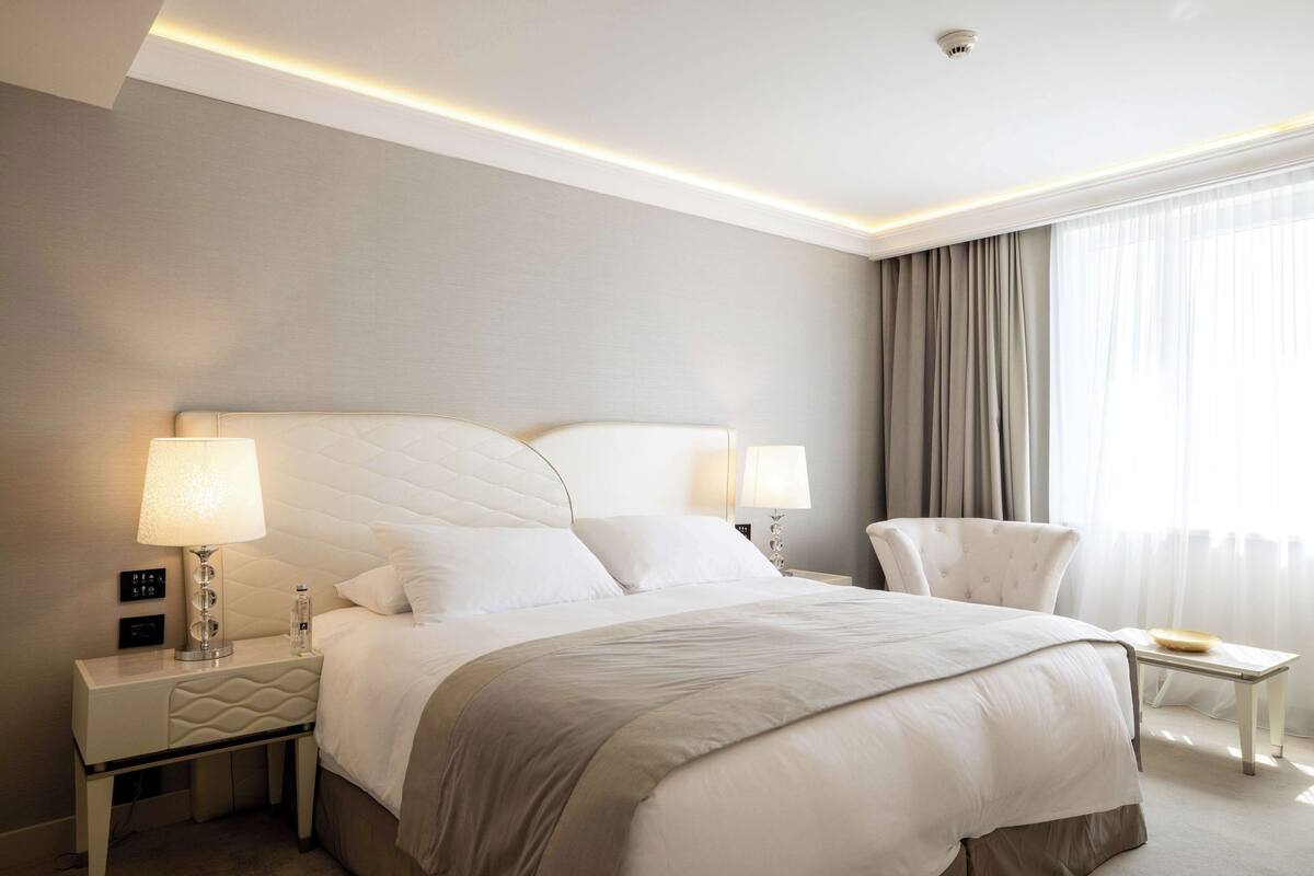 Une chambre d’hôtel moderne avec un grand lit, deux tables de chevet avec des lampes, un fauteuil et une vue sur l’extérieur à travers des rideaux légers.