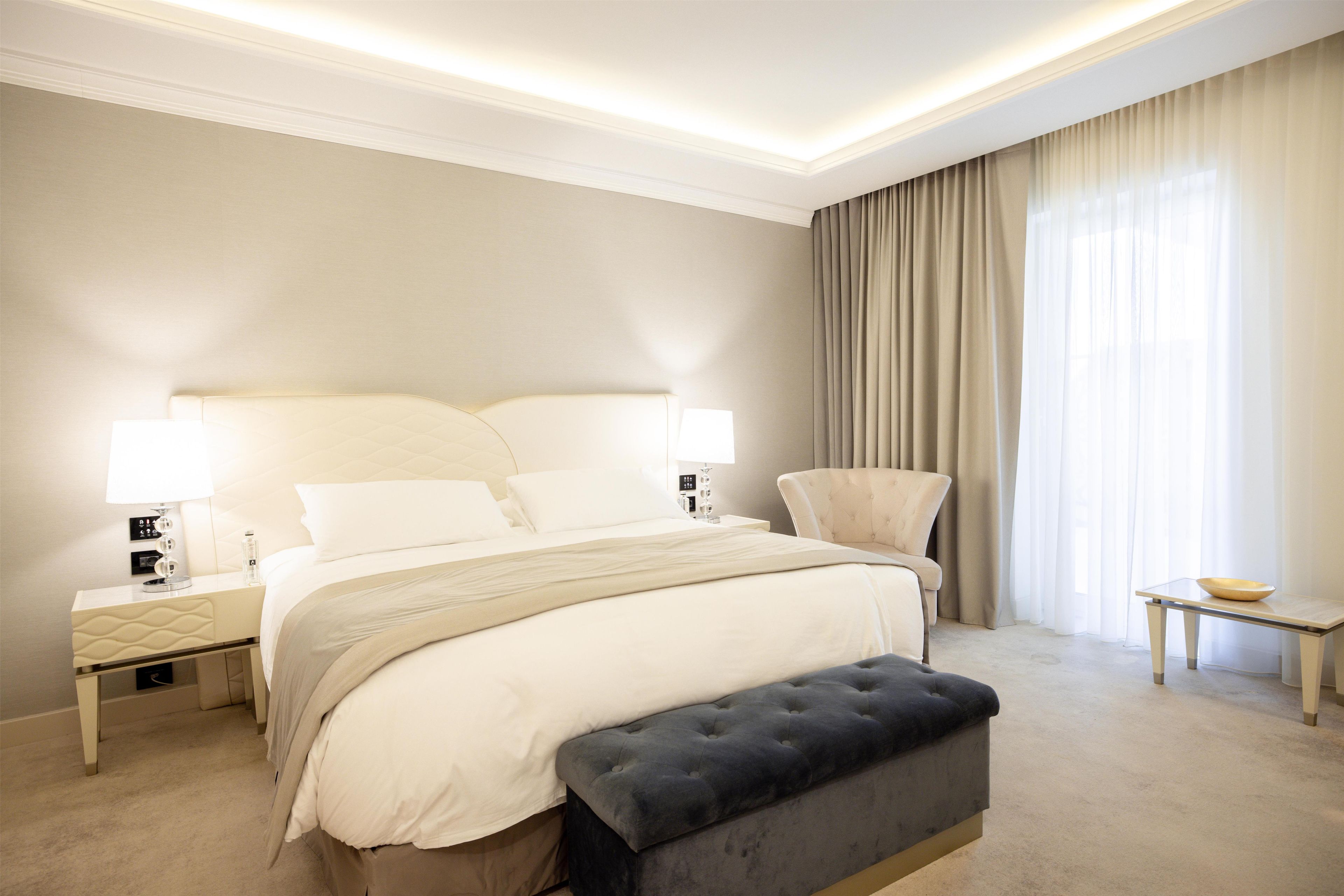 A hotel room with a large bed, bedside lamps, a chair, a small table, and a window with curtains.