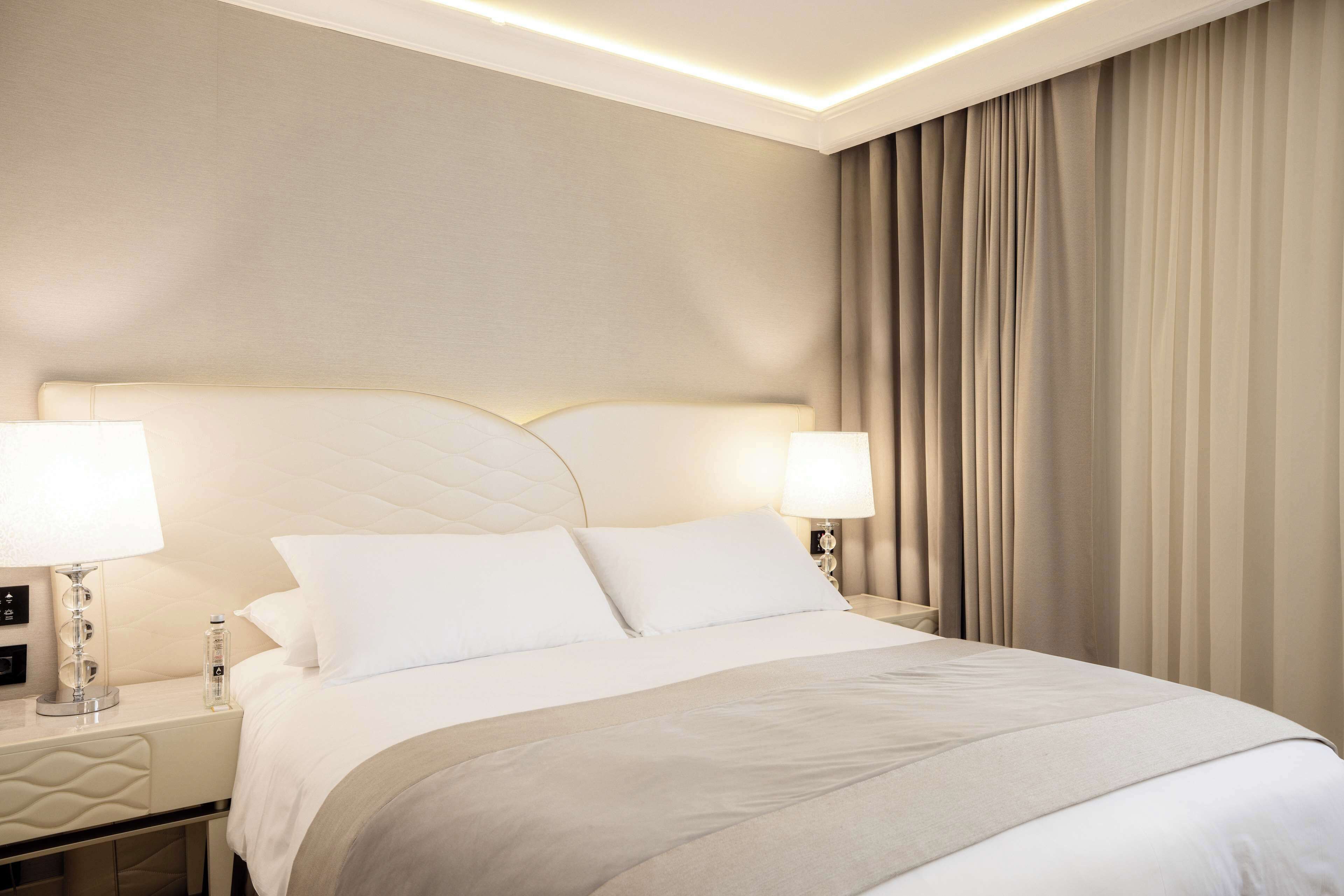 Una habitación de hotel con una cama grande, dos lámparas de noche, una mesita de noche y una cortina.