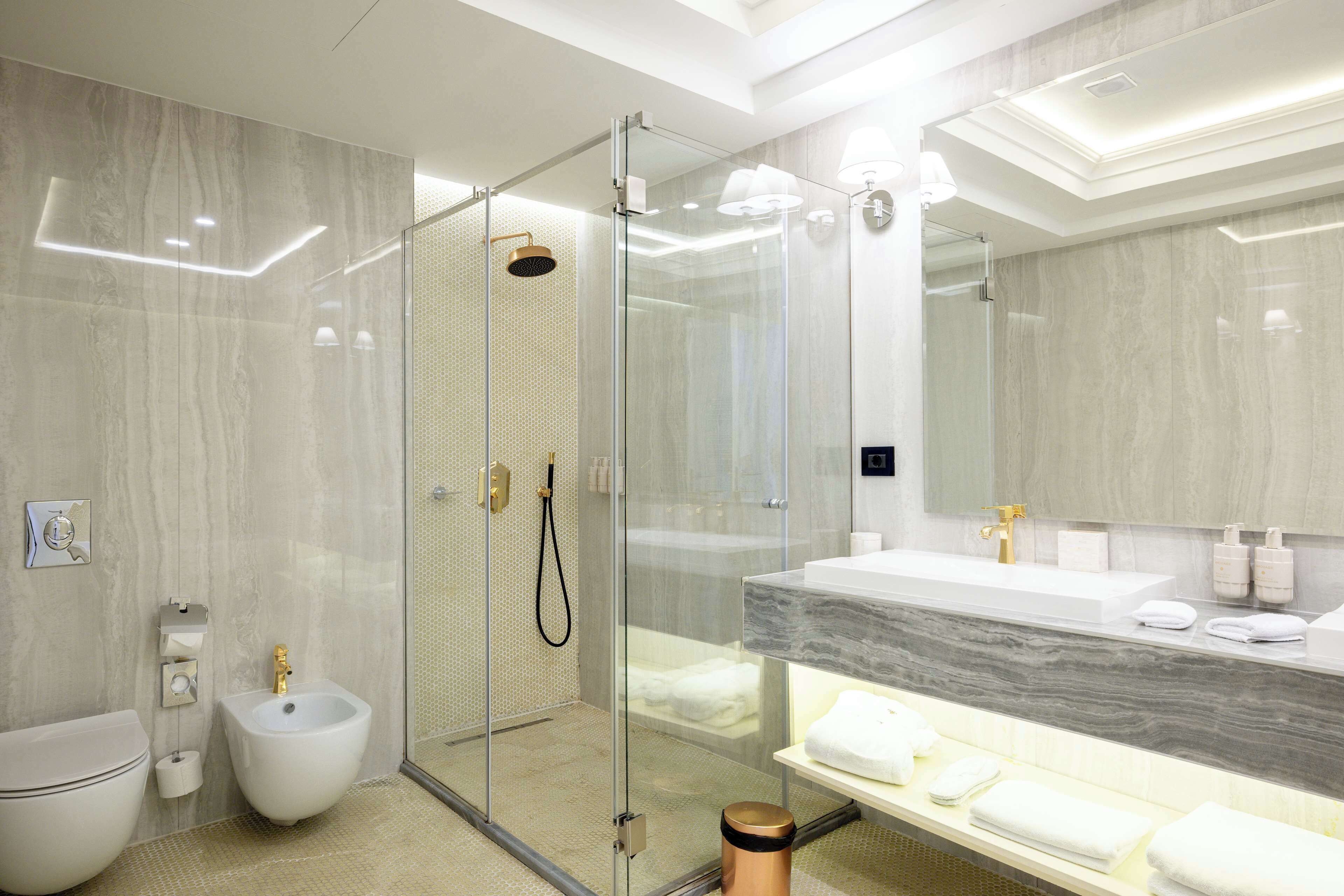 A modern bathroom with a glass-enclosed shower, a bidet, a large mirror, and a marble countertop.