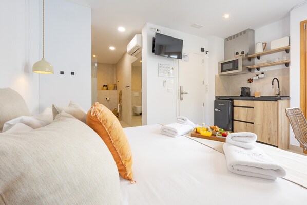 A hotel room with a bed, a TV, a kitchenette, and a bathroom visible through an open door.