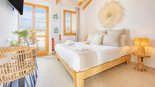 A bedroom with a bed, bedside table, lamp, wicker chair, and a view of the outdoors.