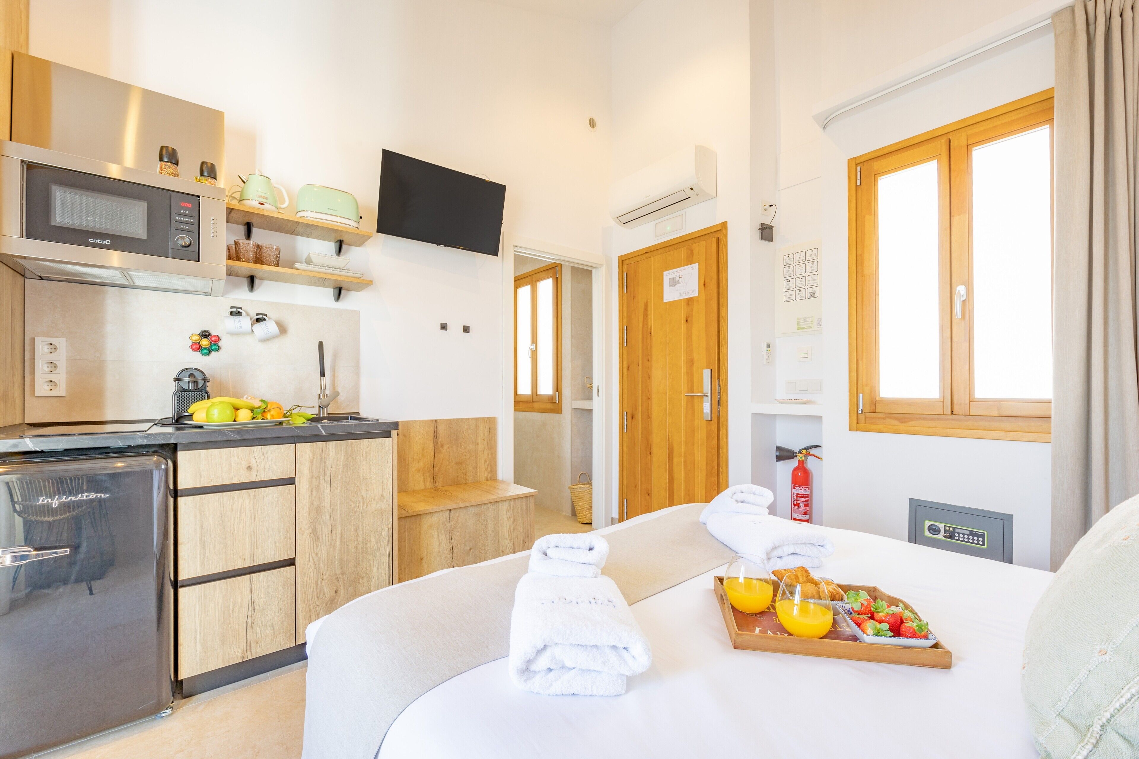 A compact hotel room with a kitchenette, a bed with towels, a breakfast tray, and a window with curtains.