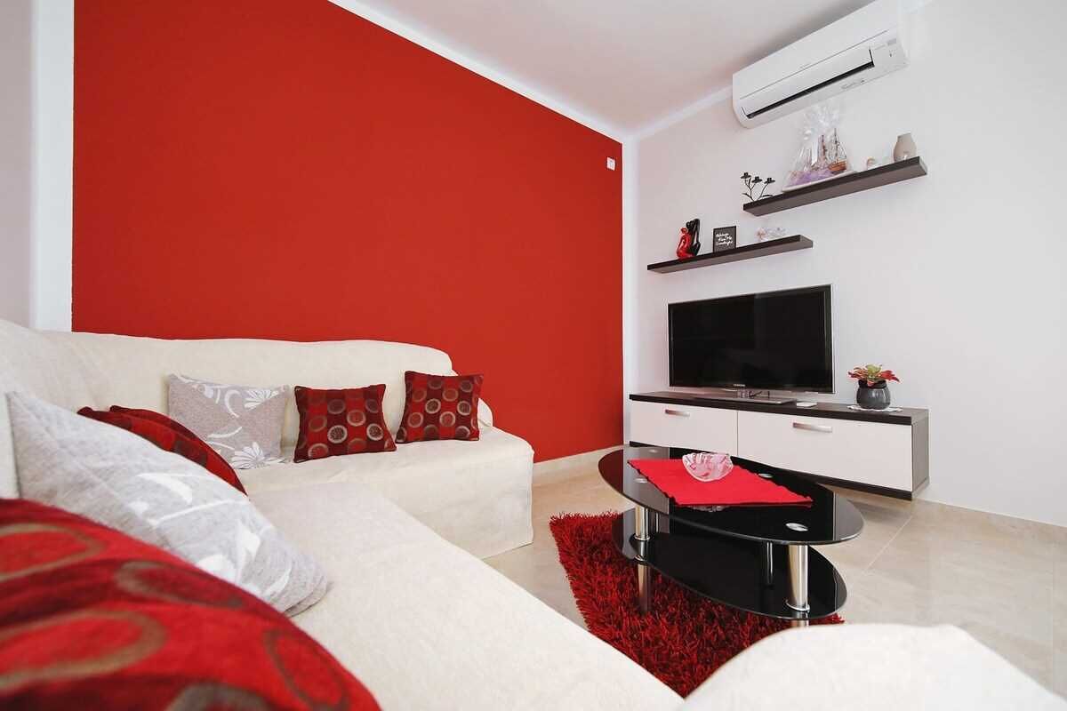 A modern living room with a red accent wall, a white sofa, a black coffee table, and a flat-screen TV on a white cabinet.