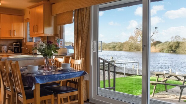 A dining area with a table set for four, a view of a lake, and a wooden deck outside. - 2 bedroom accommodation in Brundall (Brundall)