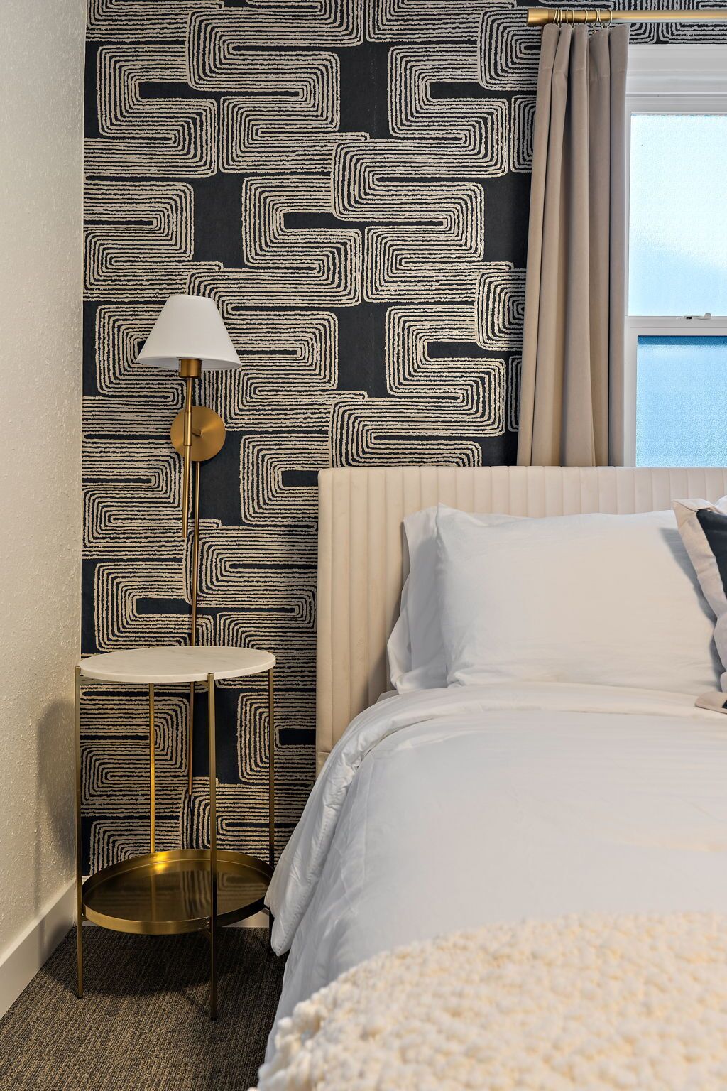 A hotel room with a patterned wall, a round table with a lamp, a bed with white linens, and a view of the sea through a window.