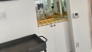 Una habitación con pared blanca, un cuadro enmarcado, un espejo que refleja una zona de cocina con encimera y cristalería, y un carro metálico negro.