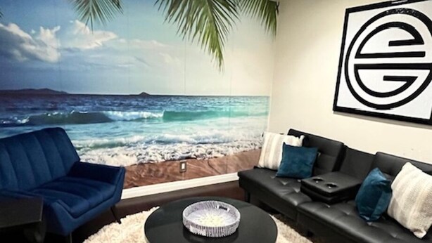 Una sala de estar con un mural de playa, un sofá negro con cojines azules, un sillón azul y una mesa de centro redonda.