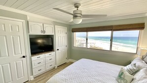 A bedroom with a bed, a TV, a ceiling fan, and a window with a beach view.