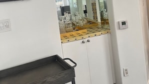 Una habitación con pared blanca, un cuadro enmarcado, un espejo que refleja una zona de cocina con encimera y cristalería, y un carro metálico negro.