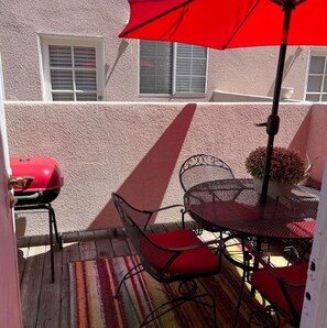 A patio with a red umbrella, a grill, and a table with chairs.
