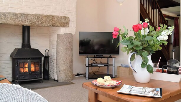 A cozy living room with a stone fireplace, a flat-screen TV, a wooden table with a vase of flowers, and a staircase in the background. - Grandma's Cottage - UK48618 (Warbstow)