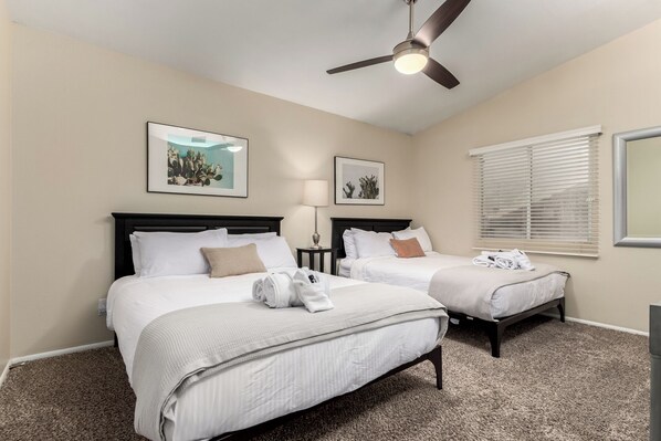 A hotel room with two beds, a ceiling fan, a window with blinds, and two framed pictures on the wall.