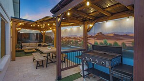 Eine Terrasse im Freien mit Grill, Holz-Pergola und einer Landschaftsdarstellung als Wandgemälde.