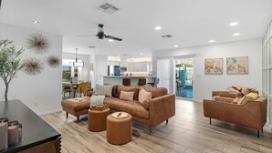 Ein geräumiges Wohnzimmer mit einem braunen Ledersofa, Poufs und einem Couchtisch. Eine Deckenlampe und Einbauleuchten sind sichtbar.