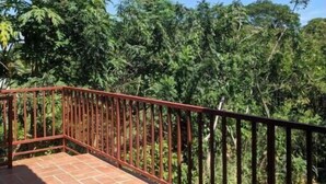 Un balcon doté d’une balustrade en métal rouge, d’un sol carrelé et d’un luxuriant feuillage verdoyant en arrière-plan.