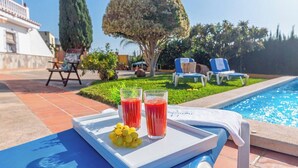 A poolside setting with a tray holding two glasses of red beverage and a bunch of grapes, a pool, and lounge chairs in the background.