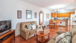 A living room with a sofa, TV, and dining area with wooden cabinets and a kitchen in the background.