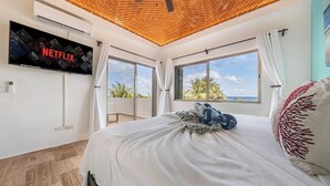 Un dormitorio con una cama grande, ventilador de techo, televisor montado en la pared y vistas a palmeras a través de las ventanas.