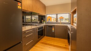 A modern kitchen with a refrigerator, stove, oven, and dishwasher. It has wooden cabinets, a window with a view of houses, and a light-colored floor.