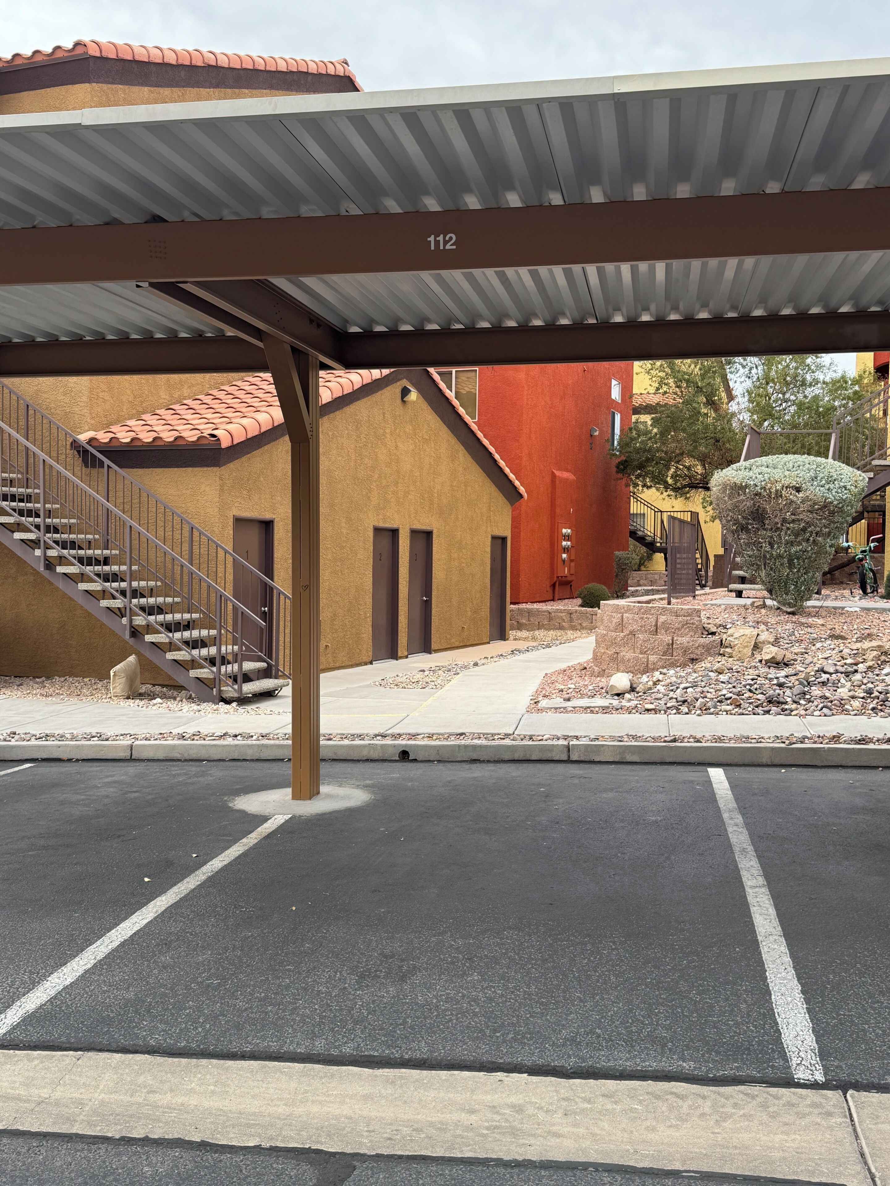 A parking lot with a covered walkway leading to a building with a red roof and beige walls, marked with the number 112.