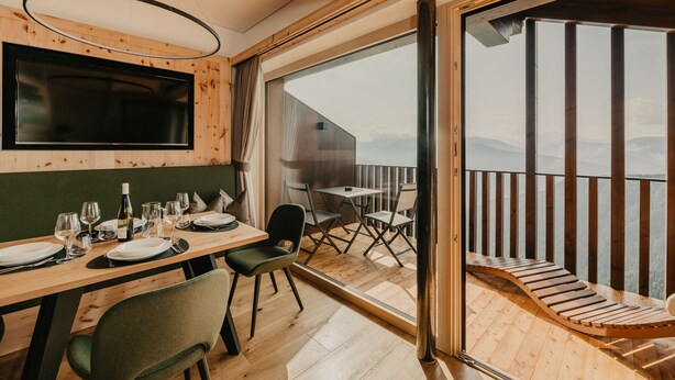 Een eethoek met een tafel voor vier personen, een aan de muur bevestigde televisie en een balkon met een houten terras en een stoel.