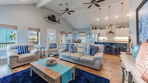A spacious living room with a beige sofa, a wooden coffee table, and a blue rug. There are ceiling fans, pendant lights, and a view of the kitchen in the background. - 4/4. Pool & Golf Cart.  1st Floor Master Suite.  2 Masters Up, Boat Parking.  
 (Port Aransas)