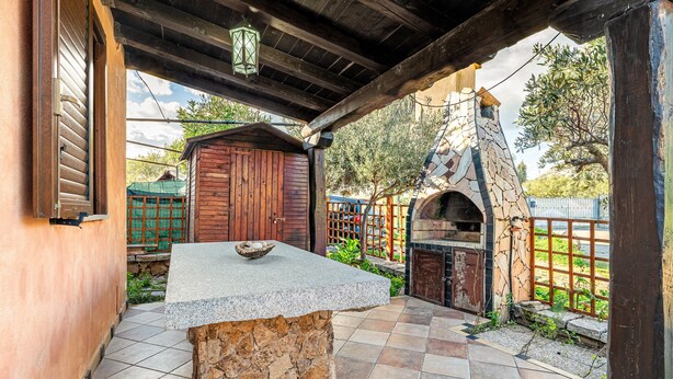 Een patio met stenen tegels, een centrale granieten tafel, een houten pergola erboven en een rustieke stenen oven.