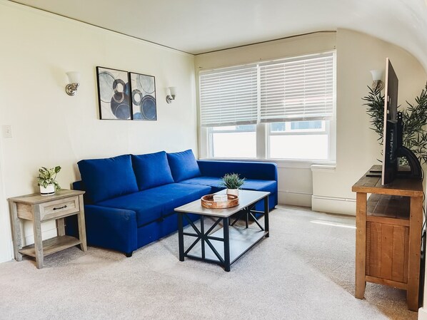 Un salon avec un canapé bleu, une table basse, un téléviseur posé sur un meuble en bois et une fenêtre avec des stores.
