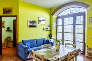 A room with a blue sofa, a dining table with chairs, a potted plant, and a window with patterned glass. - Apartment 'Stalla' with Shared Garden, Wi-Fi and Air Conditioning (Magliano in Toscana)