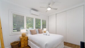Un dormitorio con una cama grande, dos mesitas de noche, un ventilador de techo y una ventana con persianas.