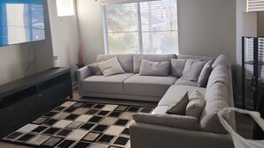 A living room with a sectional sofa, a flat-screen TV, a ceiling fan, and a large window.