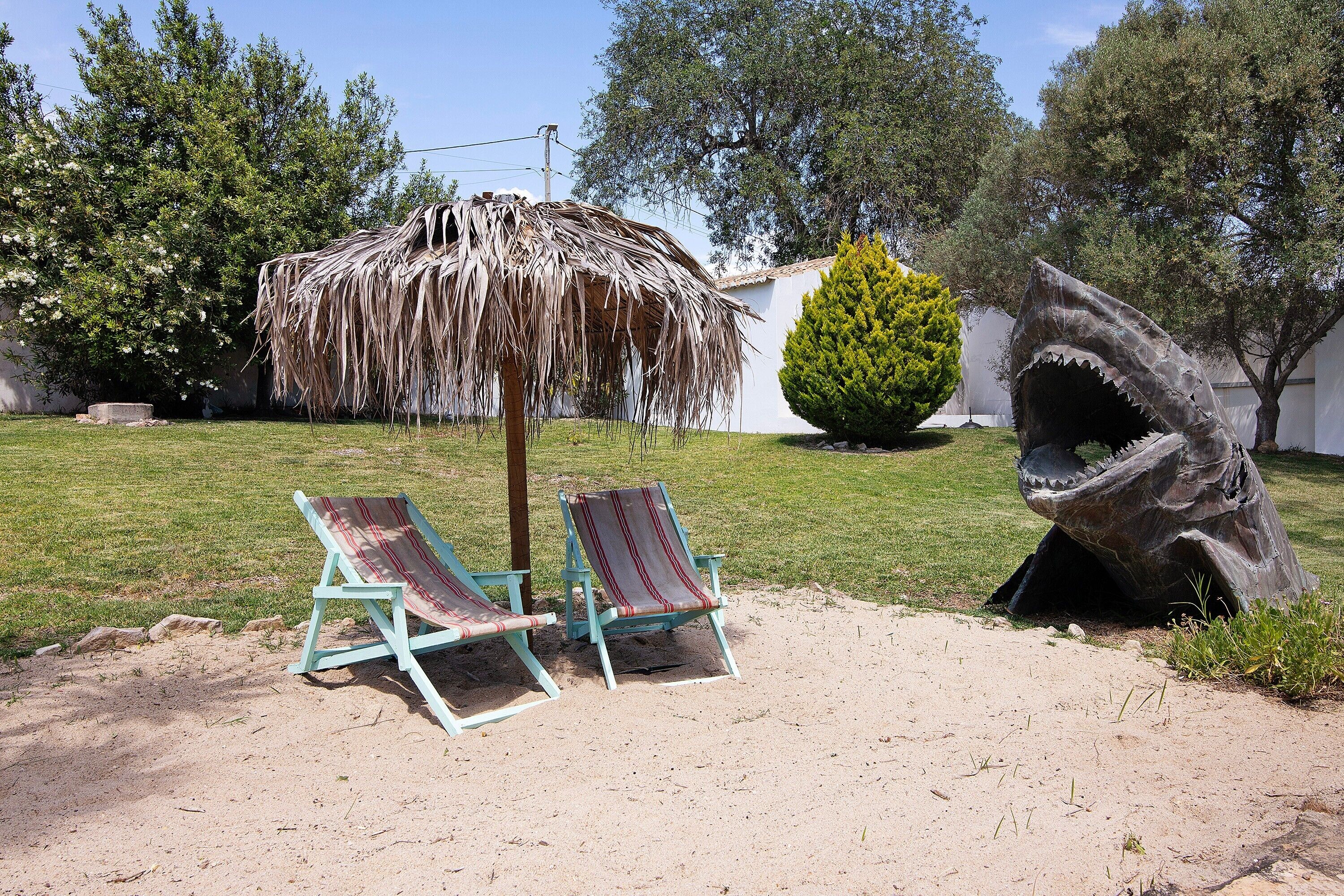 Um cenário de praia com duas cadeiras de descanso, um guarda-sol de palha e uma escultura de tubarão.