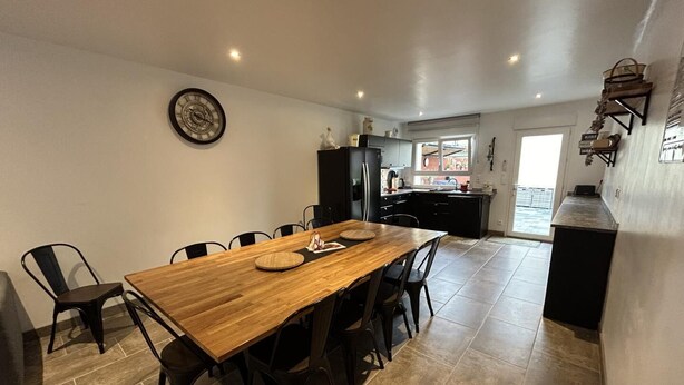 A modern kitchen with dark cabinetry, a wooden dining table, and a large window. - La Ferme Aux Deux Chevaux 1 (Francheville)