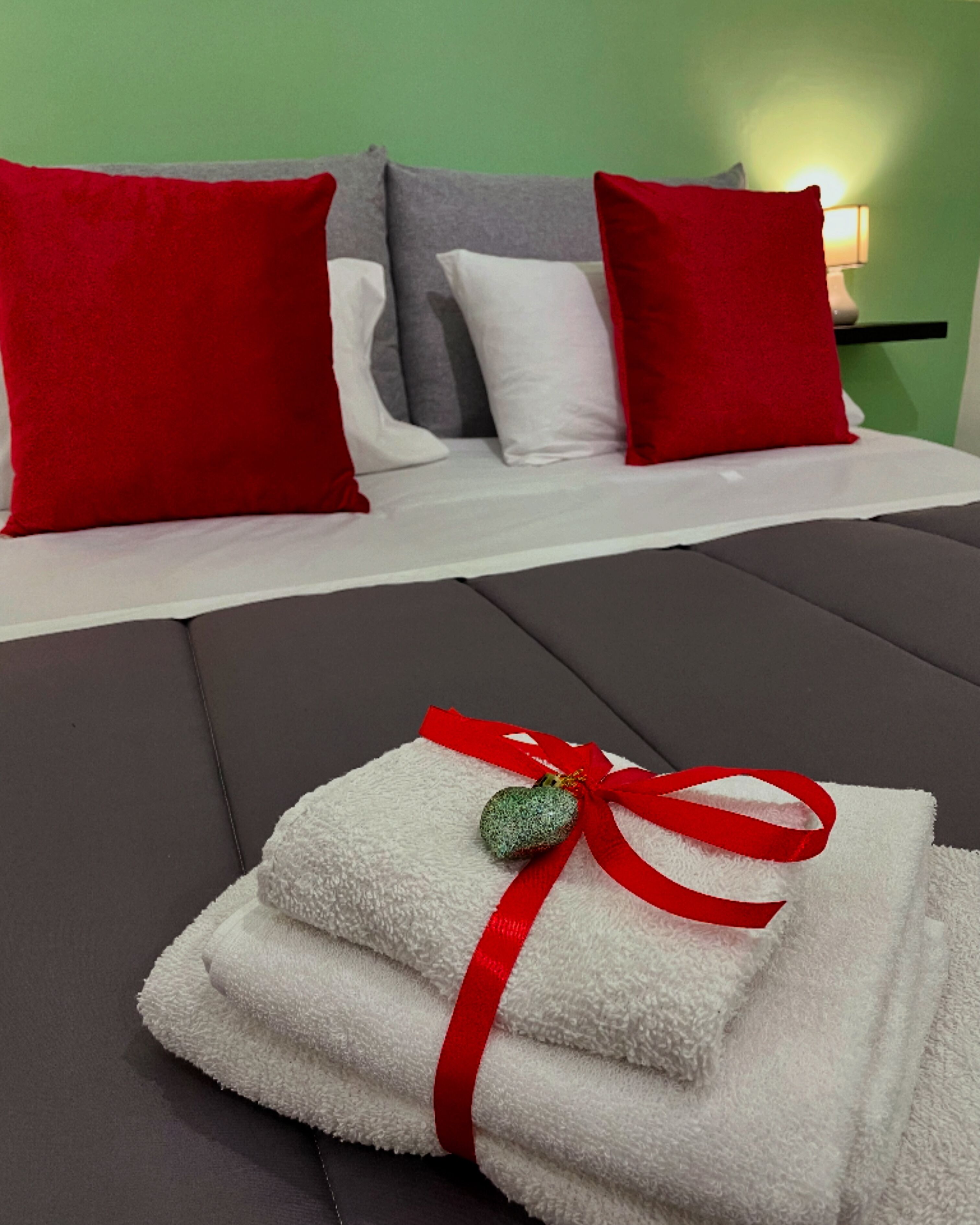 A neatly made bed with red and white pillows, a white blanket, and a red ribbon with a heart-shaped ornament on a grey mattress.