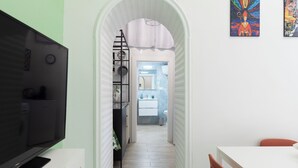 A narrow hallway leading to a bathroom with a white sink and mirror. A TV is mounted on the left wall, and there are two artworks on the right wall.