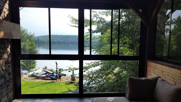 Ein Zimmer mit großem Fenster, das auf einen See und eine Wiese mit Booten blickt.