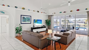A modern living room with a sofa set, a coffee table, and a TV. There is a large window with a view of a swimming pool and a building outside. - Bella Vista Villa - Waterfront Retreat with Gulf Access & Private Pool (Cape Coral)