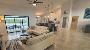 A spacious living room with a large sectional sofa, a dining area with chairs, a kitchen with modern appliances, and a view through large glass doors. - Great fall rates! Canal views! Book now! (Cape Coral)