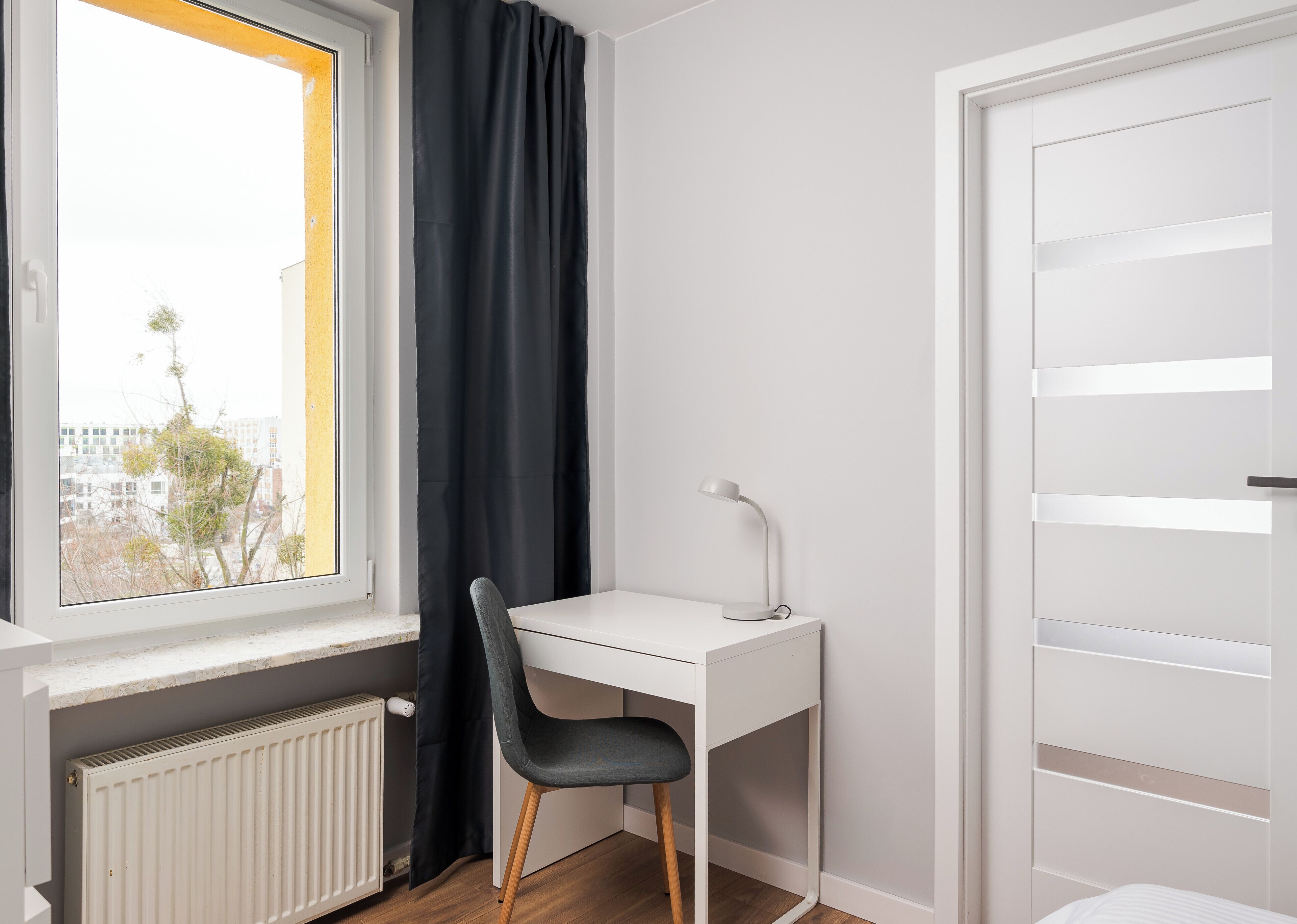 Una habitación pequeña y luminosa con un escritorio blanco, una silla negra y un ventanal con vistas a edificios y árboles.