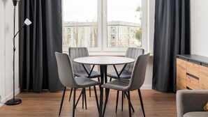 Un comedor moderno con una mesa redonda y cuatro sillas grises, una lámpara de pie negra y vistas a edificios a través de amplios ventanales.