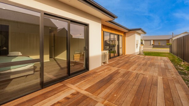 A modern house with a wooden deck, large sliding glass doors, and a visible bedroom with a bed and a chair. - Serenity Hideaway House A (Twizel)