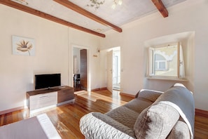 A spacious living room with wooden flooring, a sofa, a television, and a ceiling with exposed beams.