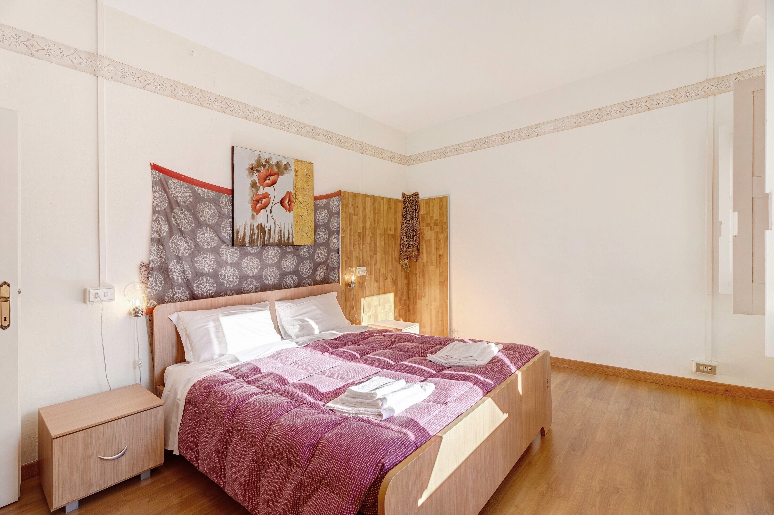 A bedroom with a wooden bed, a nightstand, a wall-mounted light, and a patterned artwork.