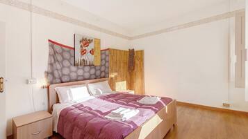 A bedroom with a wooden bed, a nightstand, a wall-mounted light, and a patterned artwork.