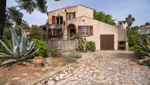 A stone-paved courtyard with a building featuring a stone facade, wooden shutters, and a large cactus plant.