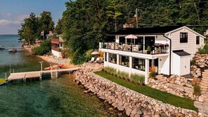 En fastighet vid vattnet med en brygga, ett stort hus med en balkong och en stenig strandlinje.