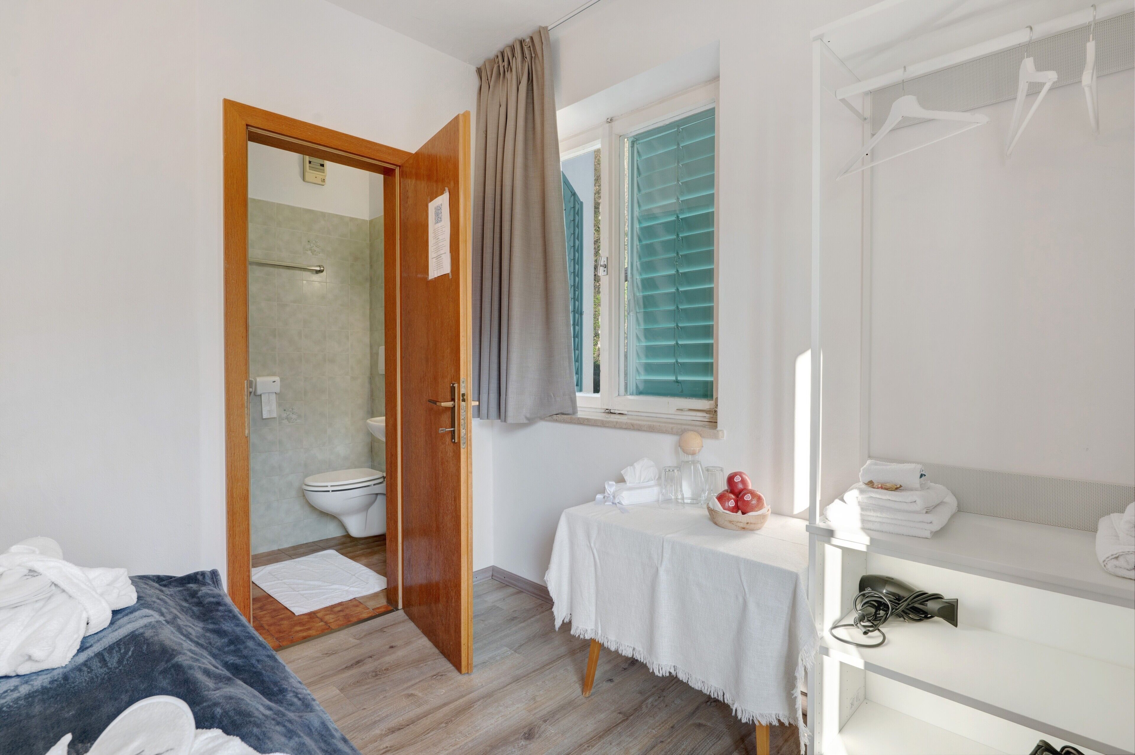 Habitació amb una porta de fusta que dona a un bany, una taula petita amb articles de tocador, un mirall i un llit amb llençols blaus.
