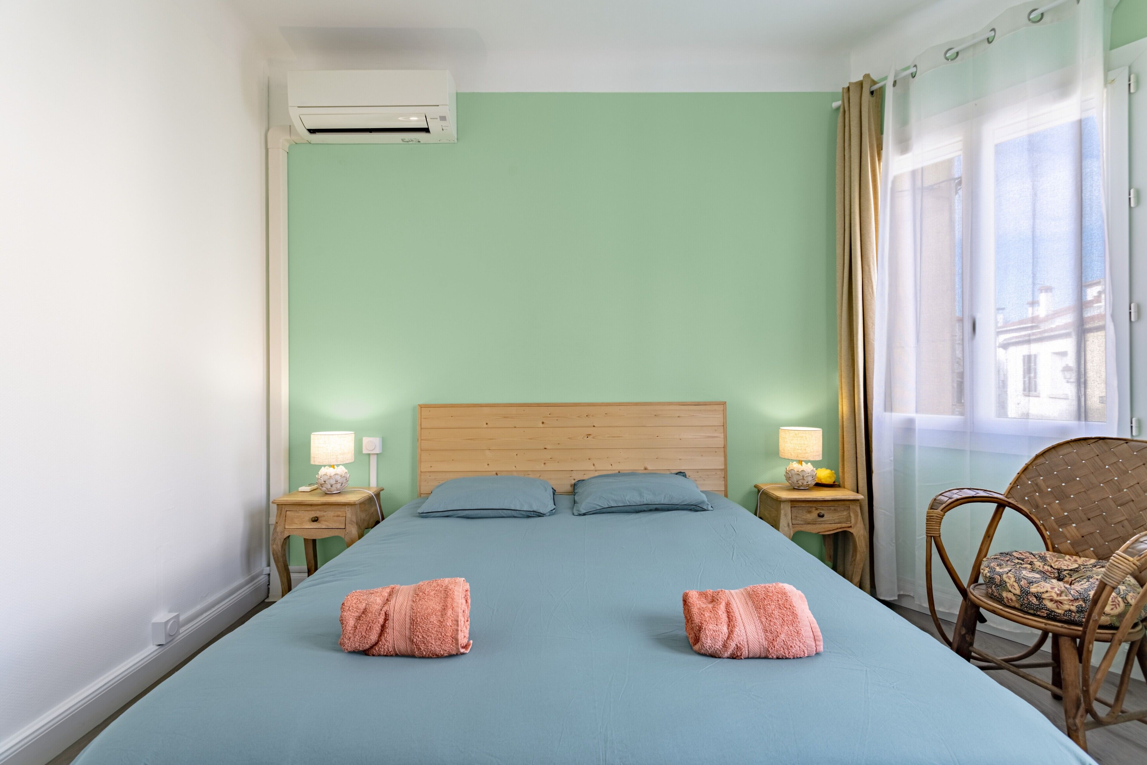Een slaapkamer met een bed, twee nachtkastjes met lampen, een raam met gordijnen en een rieten stoel.
